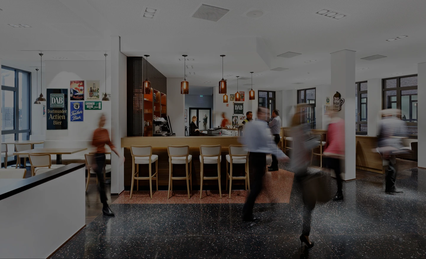 Menschen gehen durch eine moderne Bar mit Holztischen und Theke, beleuchtet durch Hängelampen.