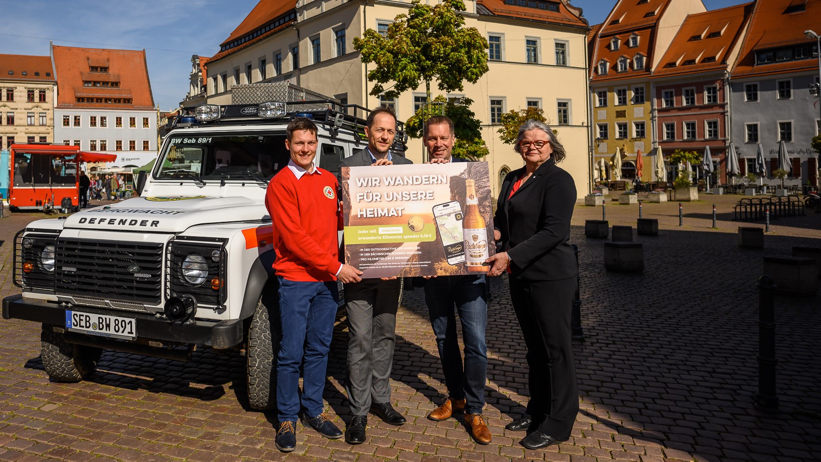Four people are standing smiling in front of an SUV, holding a large advertising sign.