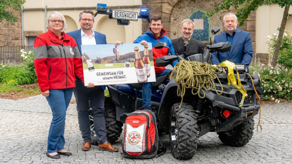 Five people are presenting a poster next to an ATV in front of a historic building.