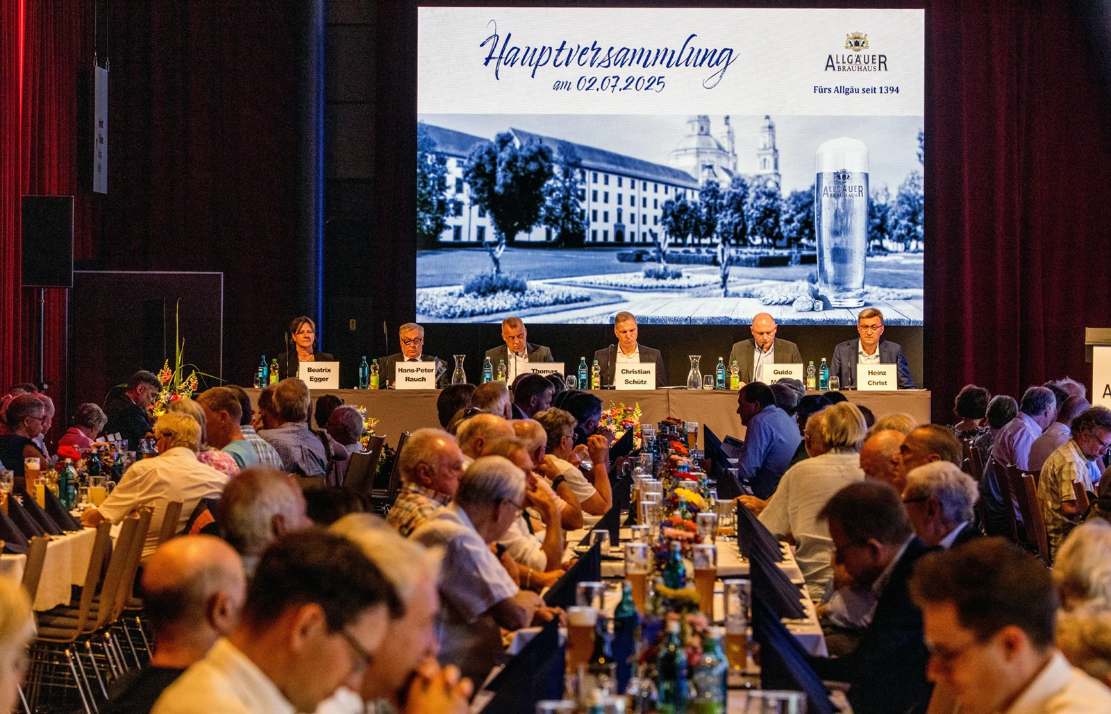 Sechs Personen sitzen auf einem Podium mit Publikum im Saal. Im Hintergrund eine Leinwand mit Datum und Firmenlogo.
