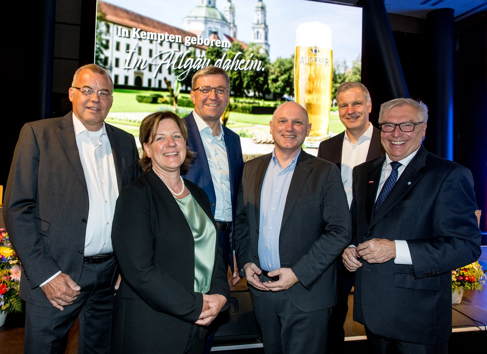 Sechs Personen in Businesskleidung stehen nebeneinander und lächeln in die Kamera. Im Hintergrund ein Allgäuer Brauhaus Motiv.