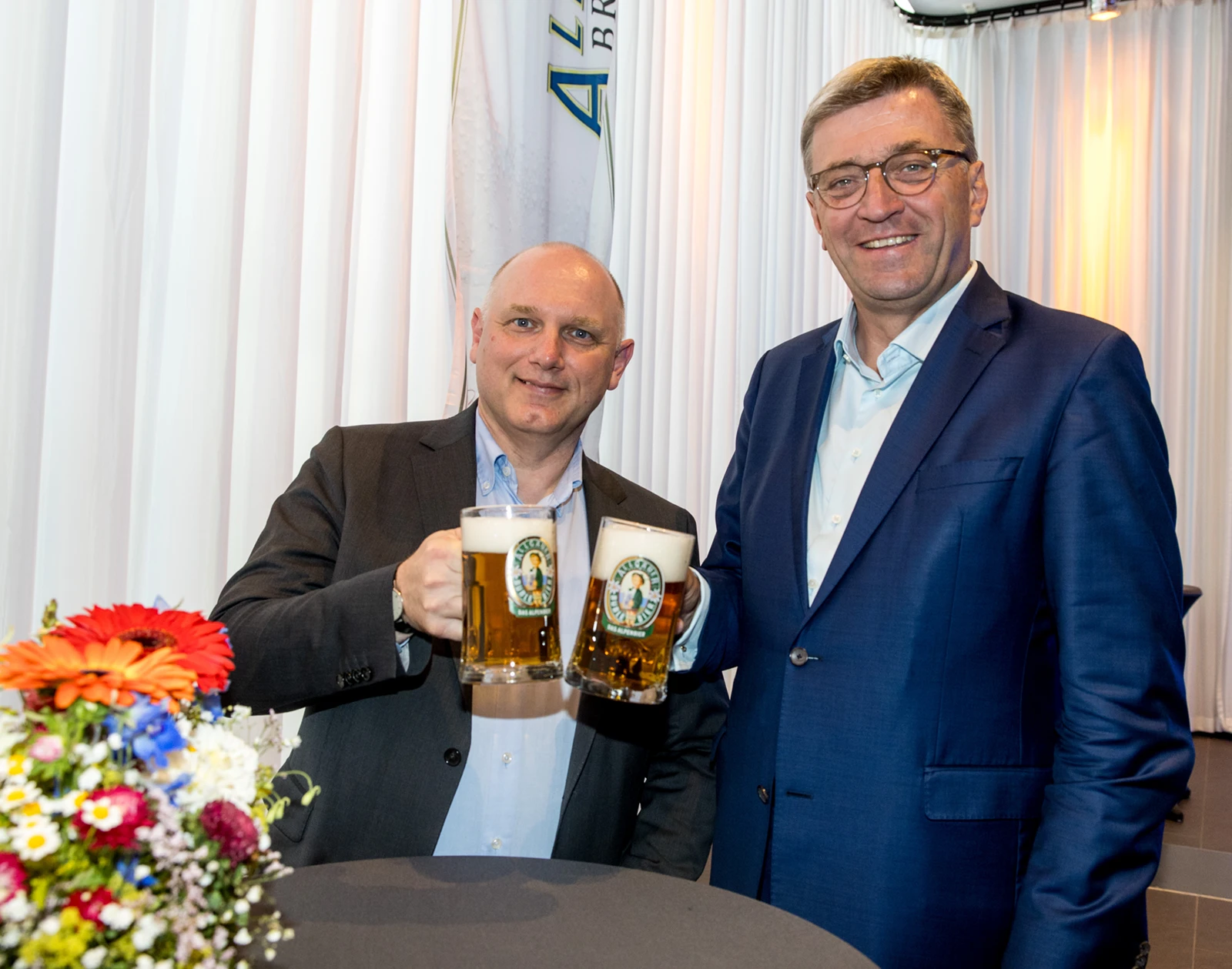 Zwei Männer im Anzug stoßen mit vollen Biergläsern an einem Stehtisch an.