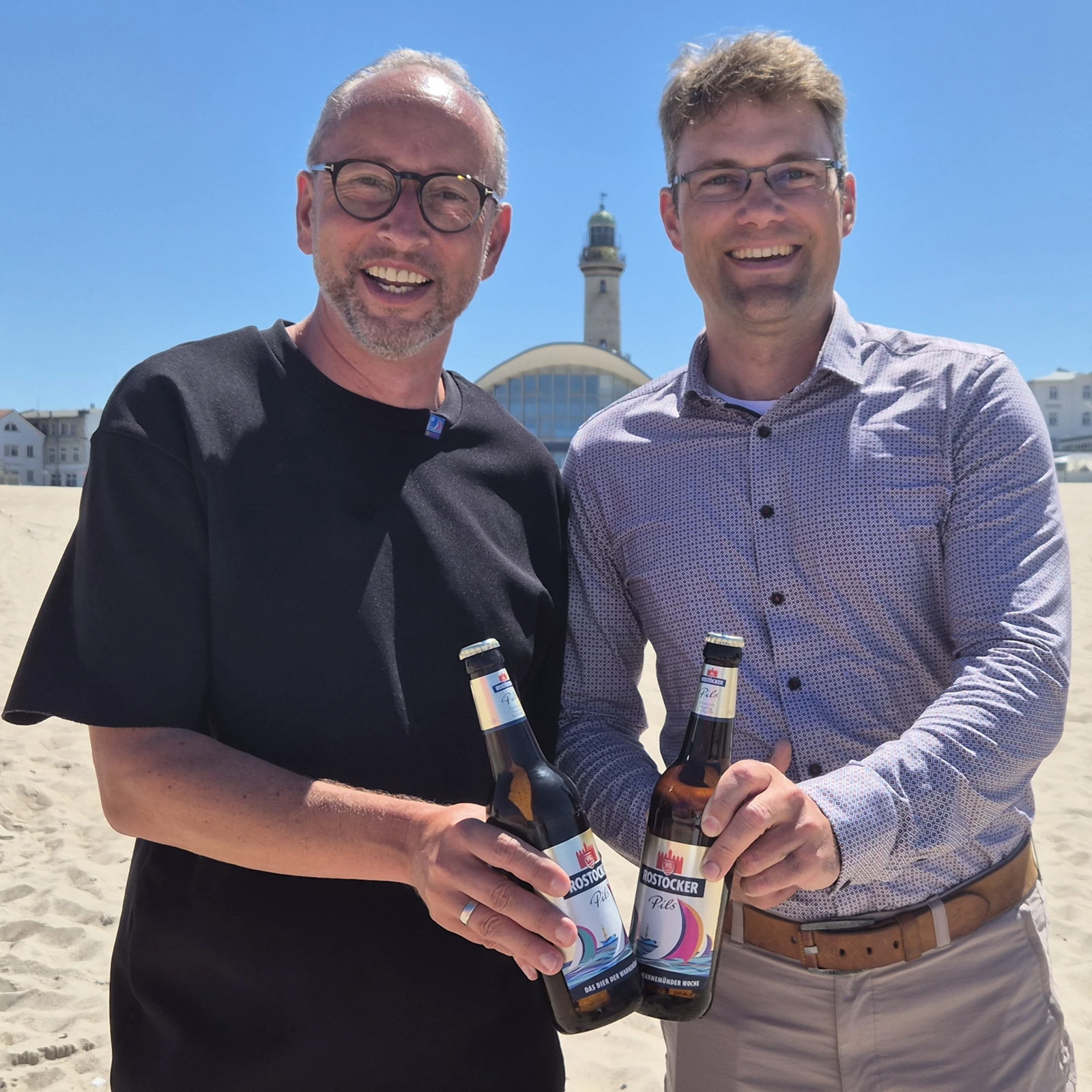 Zwei Männer stehen im Sand nebeneinander und halten die limitierte Rostocker Sondereditions-Bierflasche in der Hand.