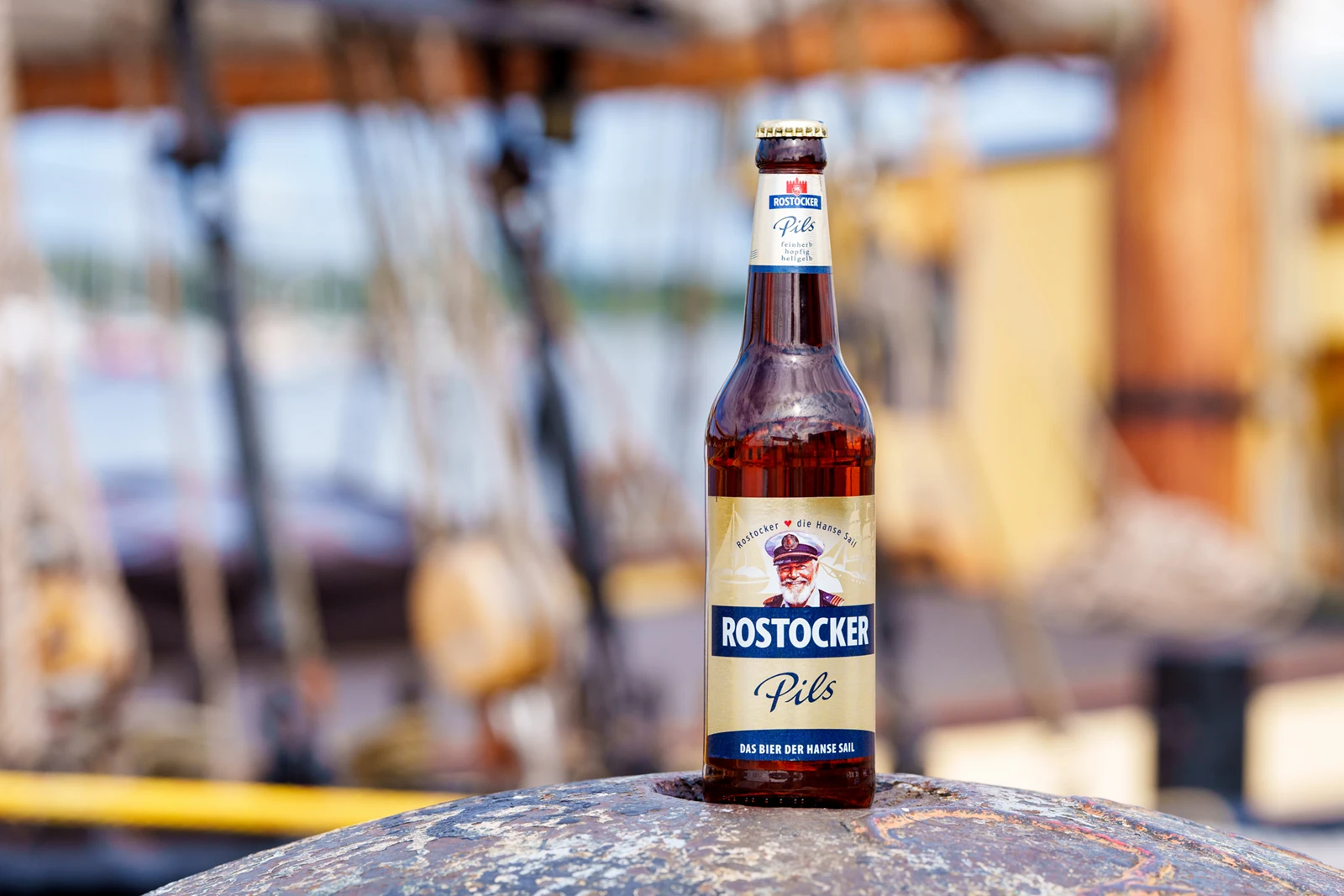 Bierflasche mit der Aufschrift „Rostocker Pils“ steht auf einem Poller. Im Hintergrund sind Teile eines Segelschiffs sichtbar.