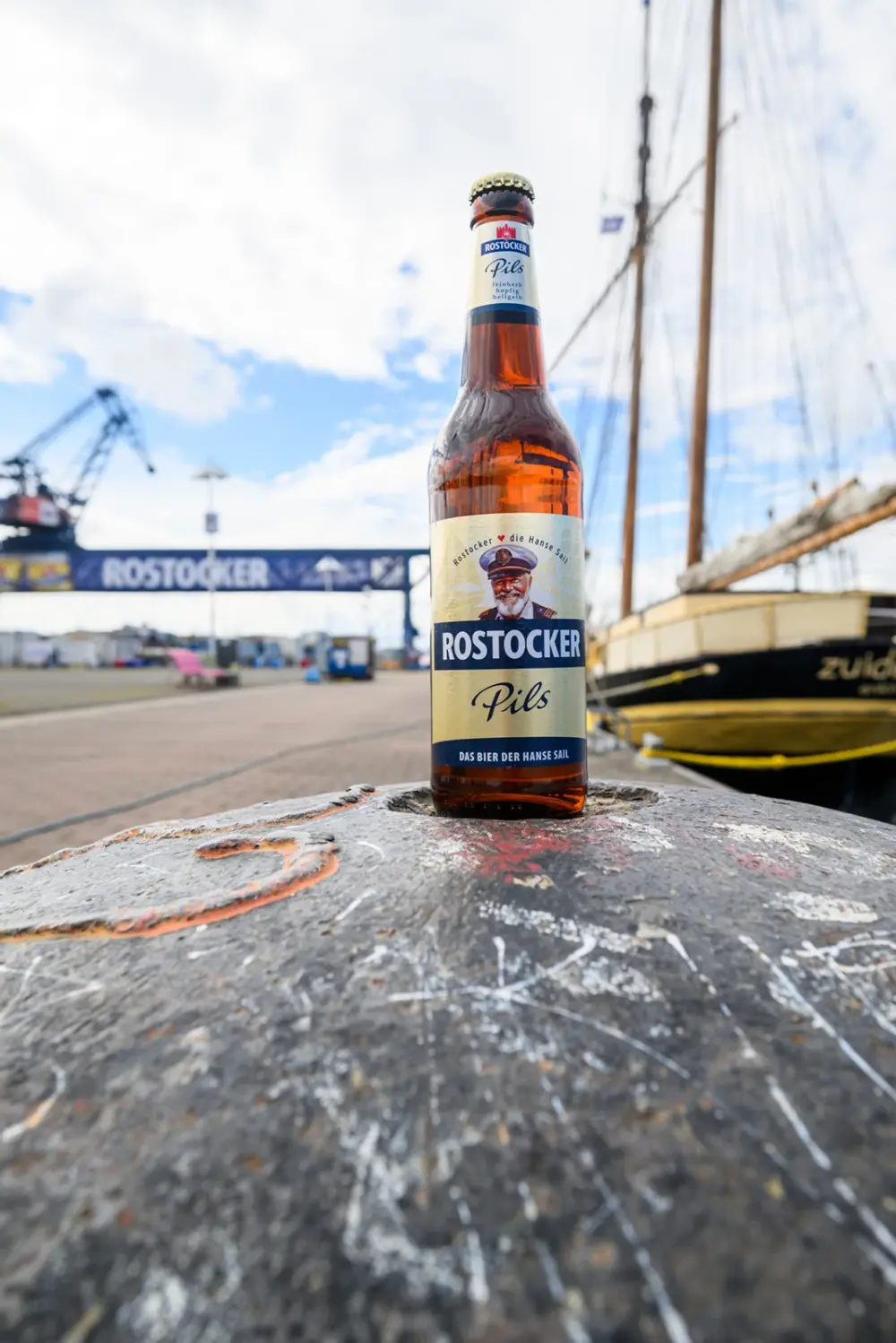 Braune Bierflasche mit dem Etikett „Rostocker Pils“ steht auf einem Poller. Im Hintergrund ein Segelschiff und ein Hafenkran.