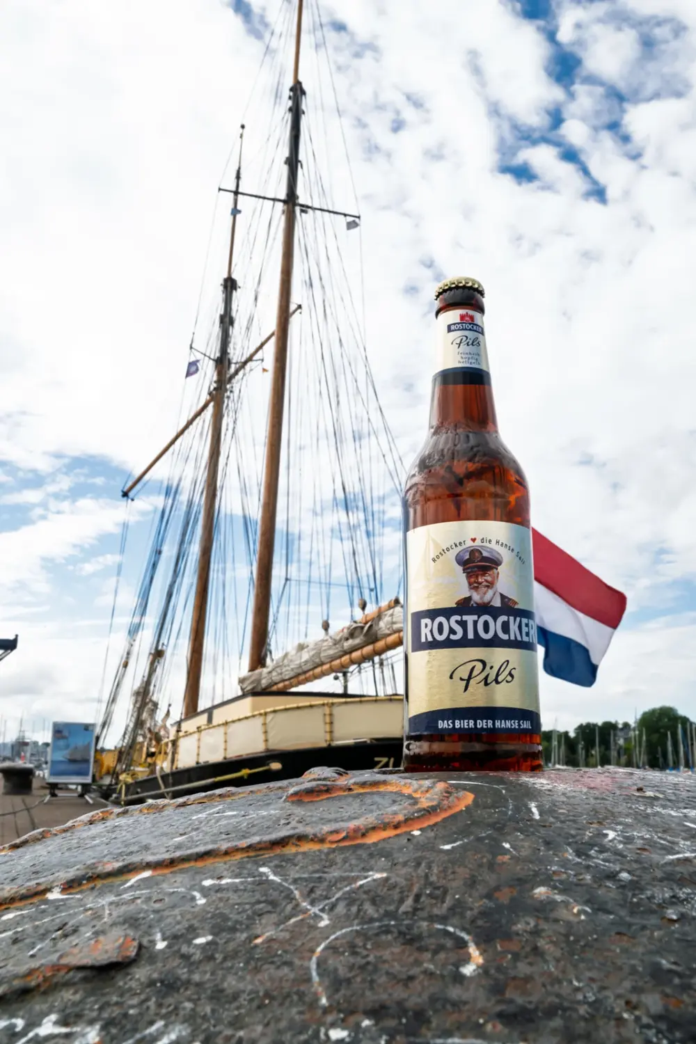 Braune Bierflasche mit der Aufschrift „Rostocker Pils“ steht auf einem Poller. Im Hintergrund ein Segelschiff mit niederländischer Flagge.
