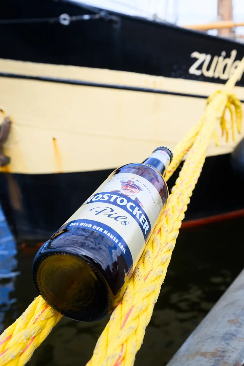 Bierflasche mit dem Etikett „Rostocker Pils“ liegt auf einem gelben Seil. Im Hintergrund ist ein schwarzes Schiff mit Schriftzug sichtbar.