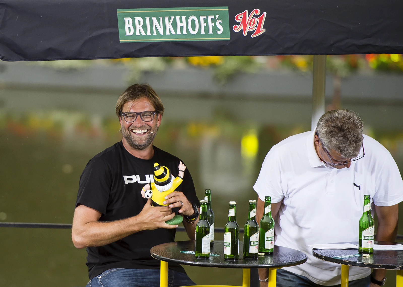 Lachender Mann hält eine kleine Figur in Fußballkleidung, neben ihm sitzt ein Mann an einem Tisch mit Brinkhoffs Bierflaschen.