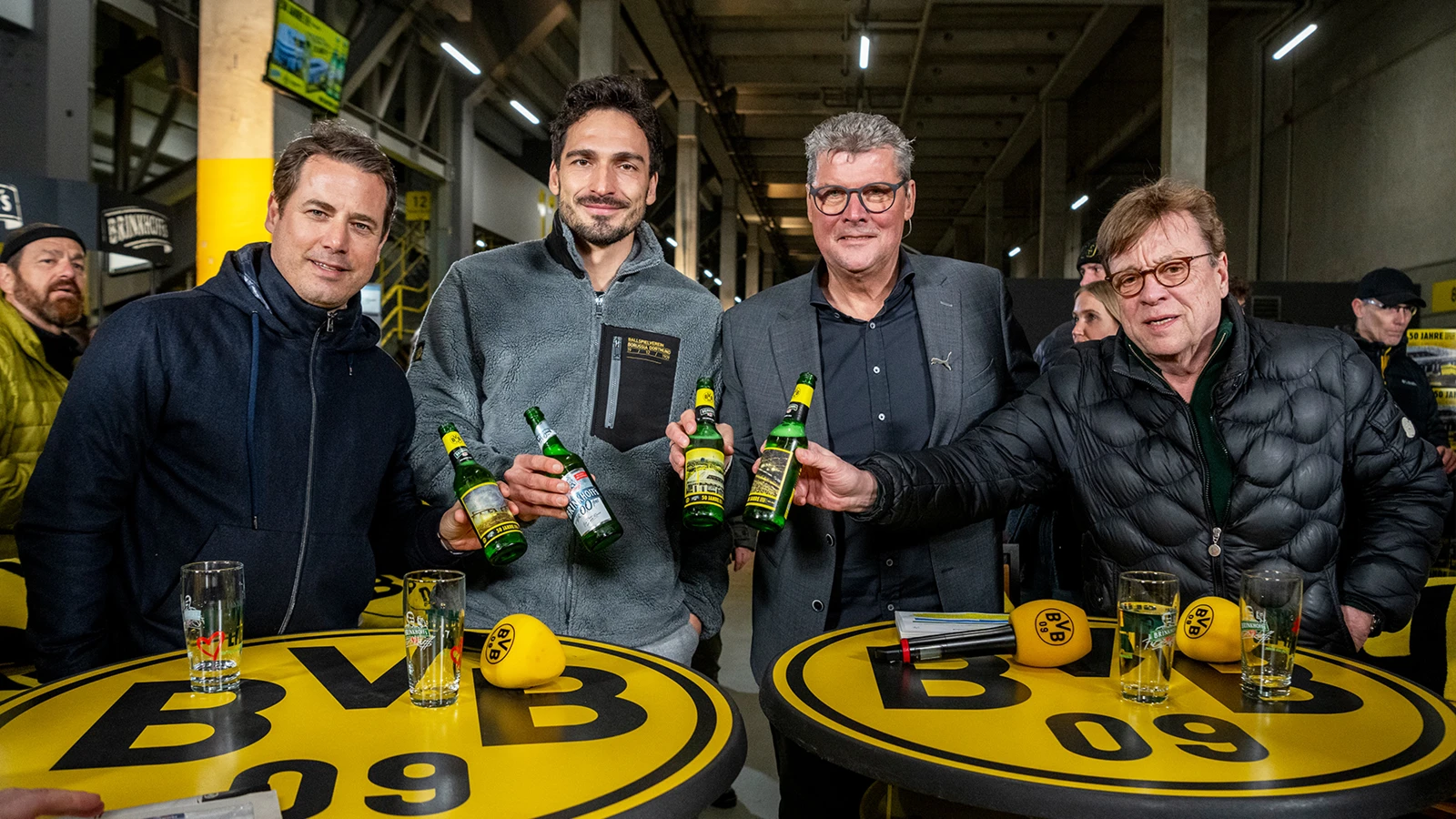 Vier Männer halten Brinkoffs Bierflaschen und stehen an gelben Tischen mit BVB Logo in einer Stadionumgebung.