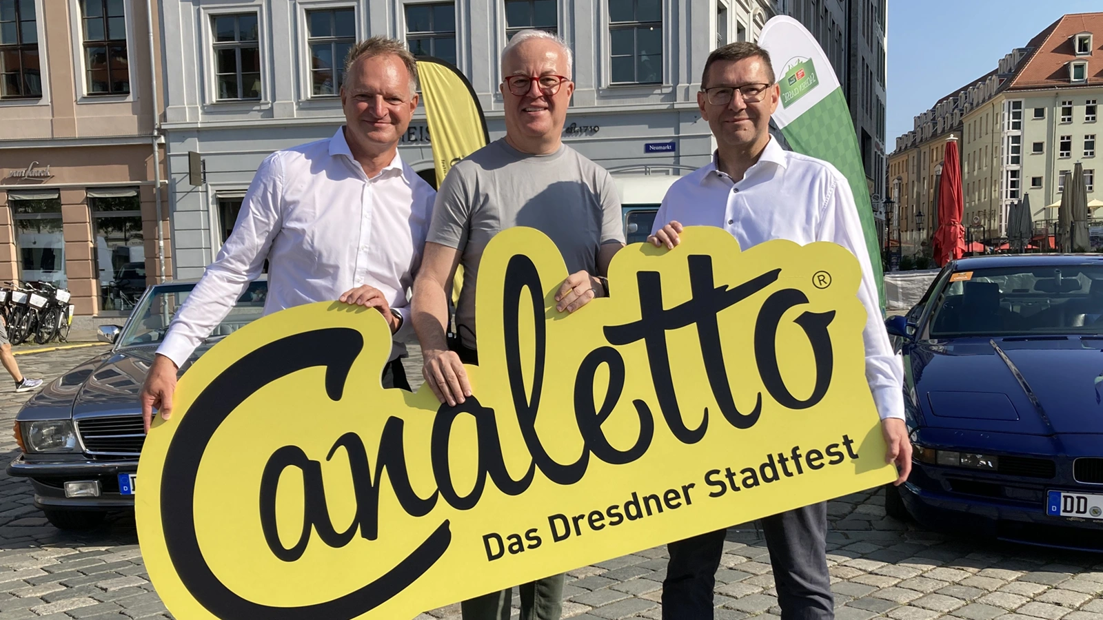 Drei Männer halten ein gelbes Schild mit der Aufschrift Canaletto Das Dresdner Stadtfest. Im Hintergrund Autos und Häuser.