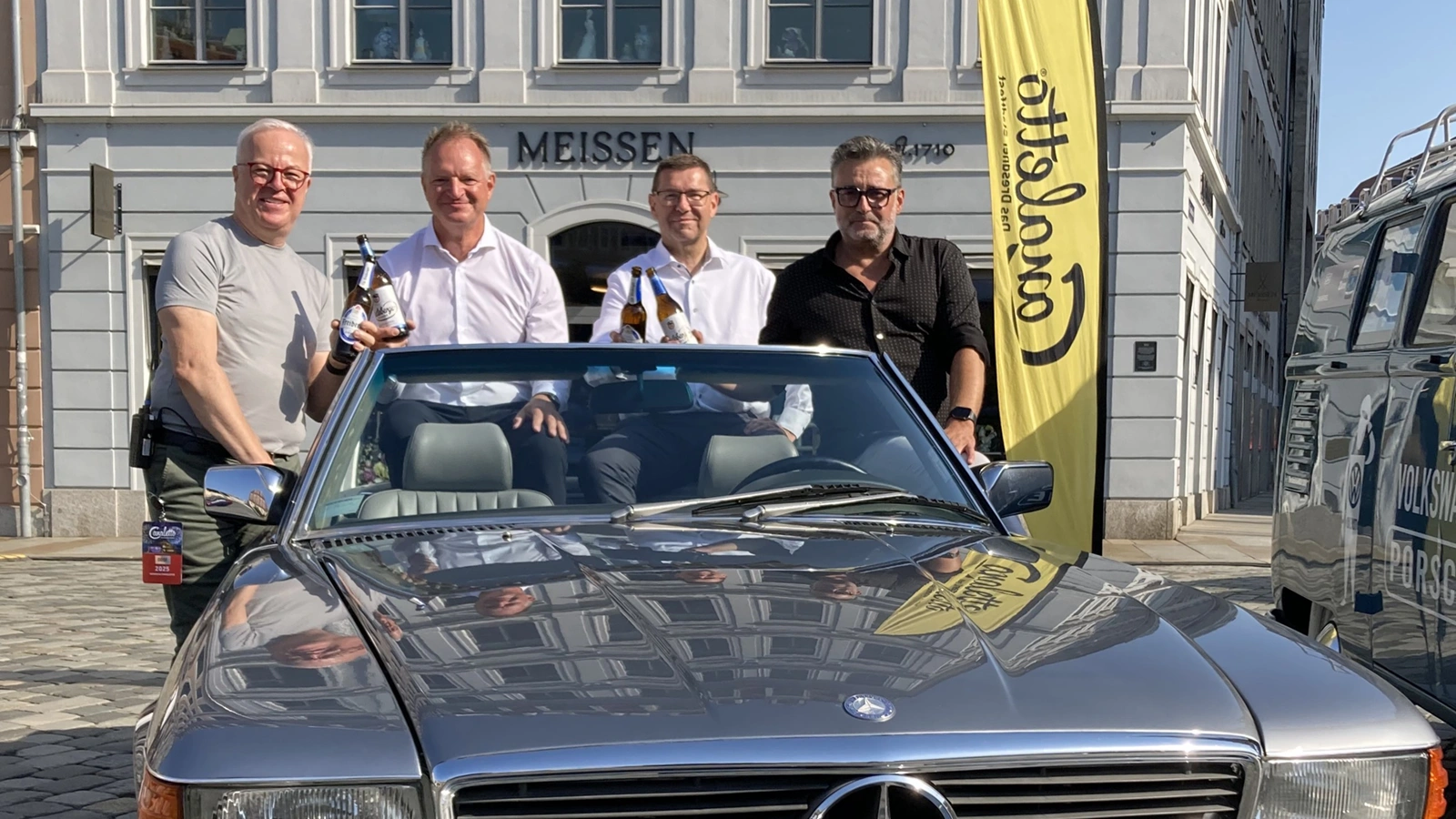 Vier Männer posieren in einem Cabrio und halten Radeberger Bierflaschen. Im Hintergrund eine Fahne mit Canaletto Aufschrift.