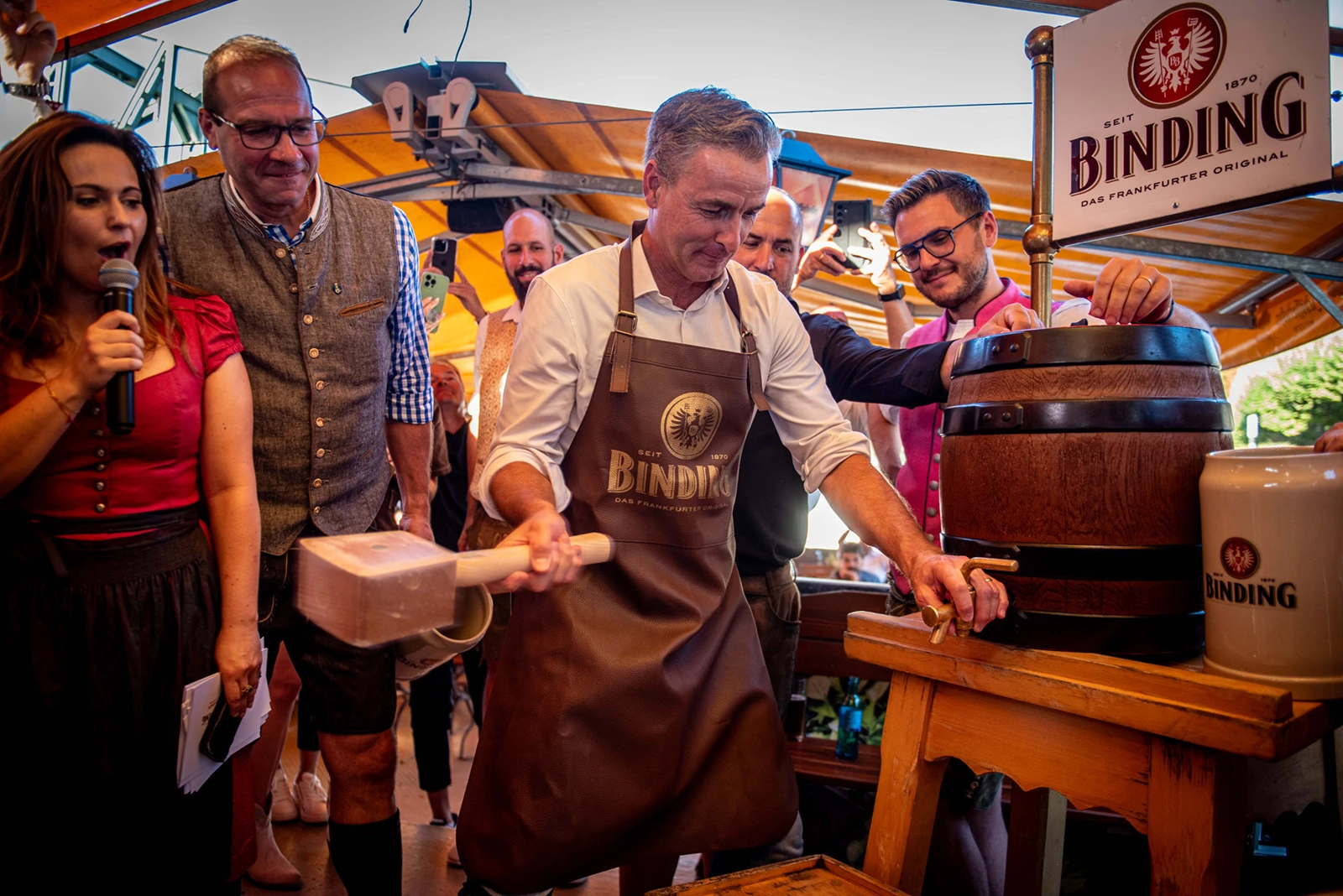 Mann schlägt mit Holzhammer Zapfhahn in Binding Bierfass, mehrere Personen stehen daneben. Im Hintergrund Zelt mit Publikum.