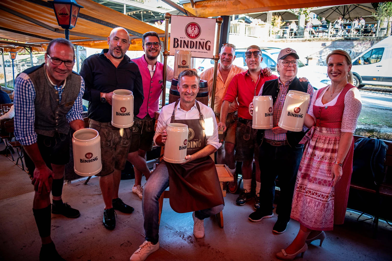 Gruppe von Menschen in Tracht hält Bierkrüge mit Binding Logo. Im Hintergrund Außengastronomie mit Sonnenschirmen.