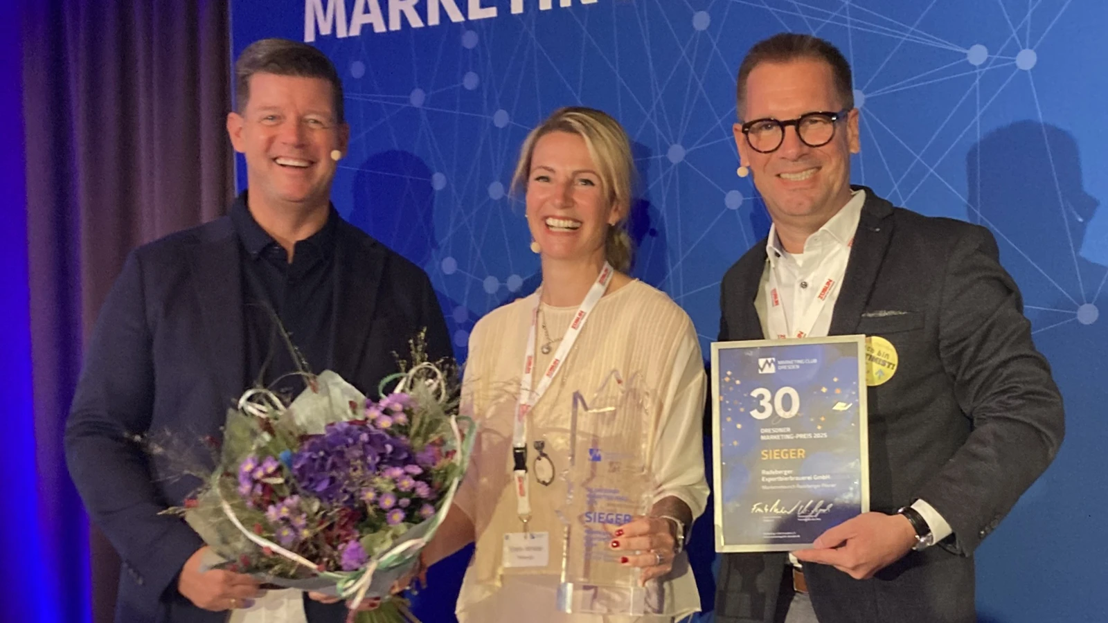 Drei Personen präsentieren Blumenstrauß, Urkunde und gläserne Trophäe bei der 30. Dresdner Marketing-Preisverleihung 2025.