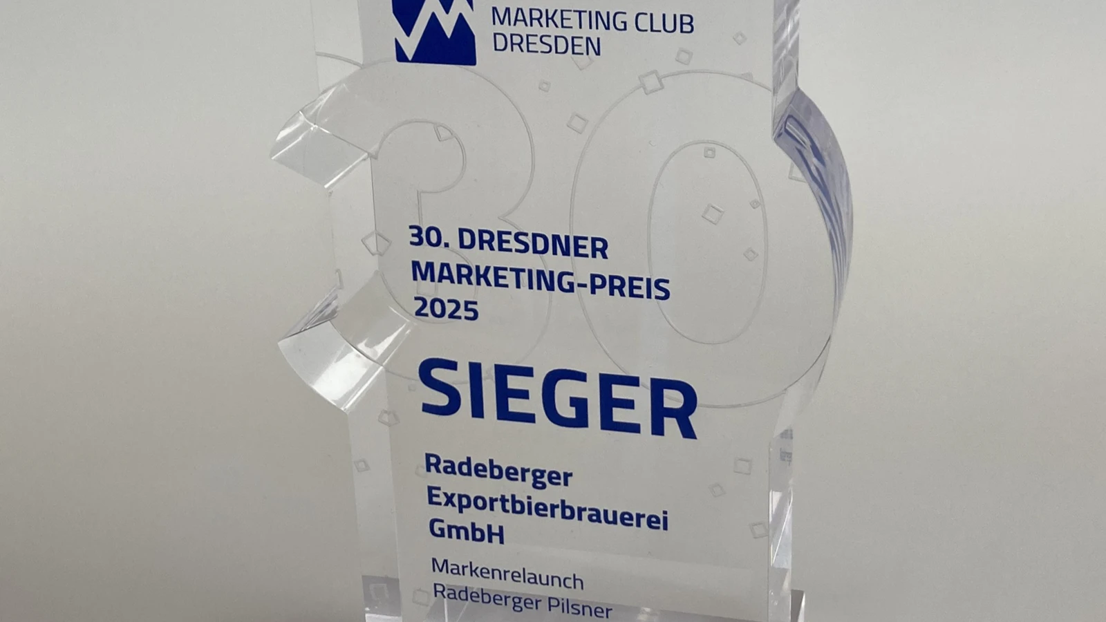 Gläserne Trophäe mit blauer Schrift und Logo des Marketing Club Dresden. Text weist auf den Dresdner Marketing Preis 2025 hin.