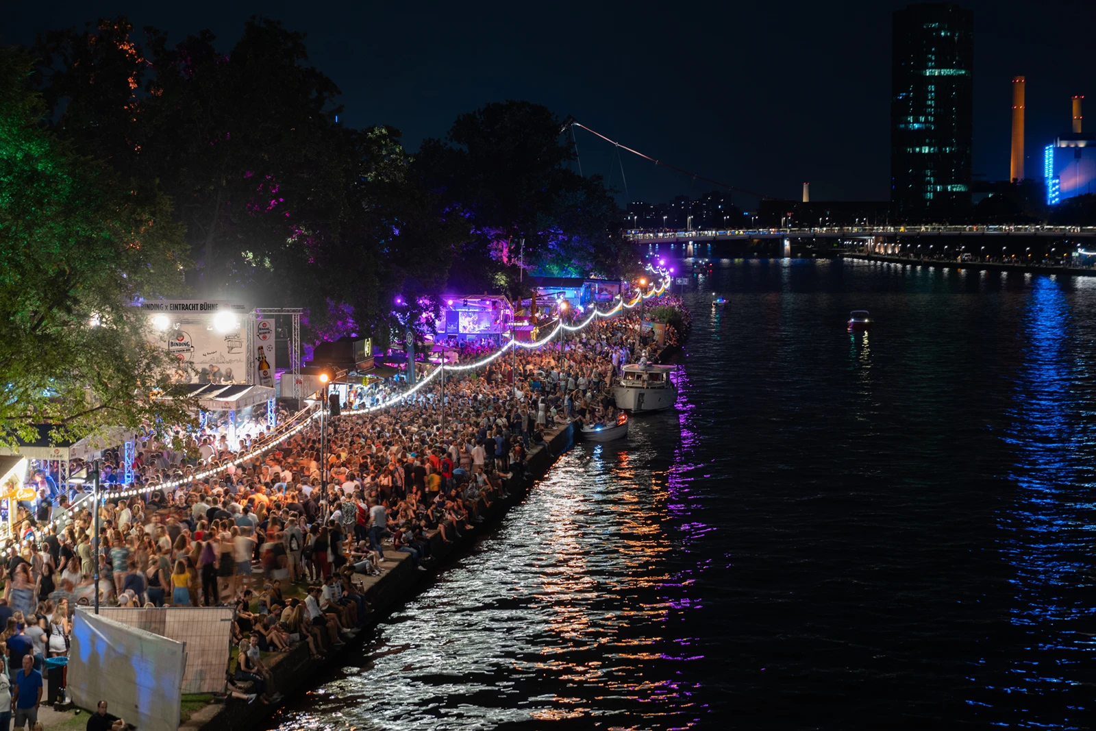 Große Menschenmenge steht abends am Museumsufer in Frankfurt. Im Hintergrund leuchten bunte Lichter, die sich im Wasser spiegeln.