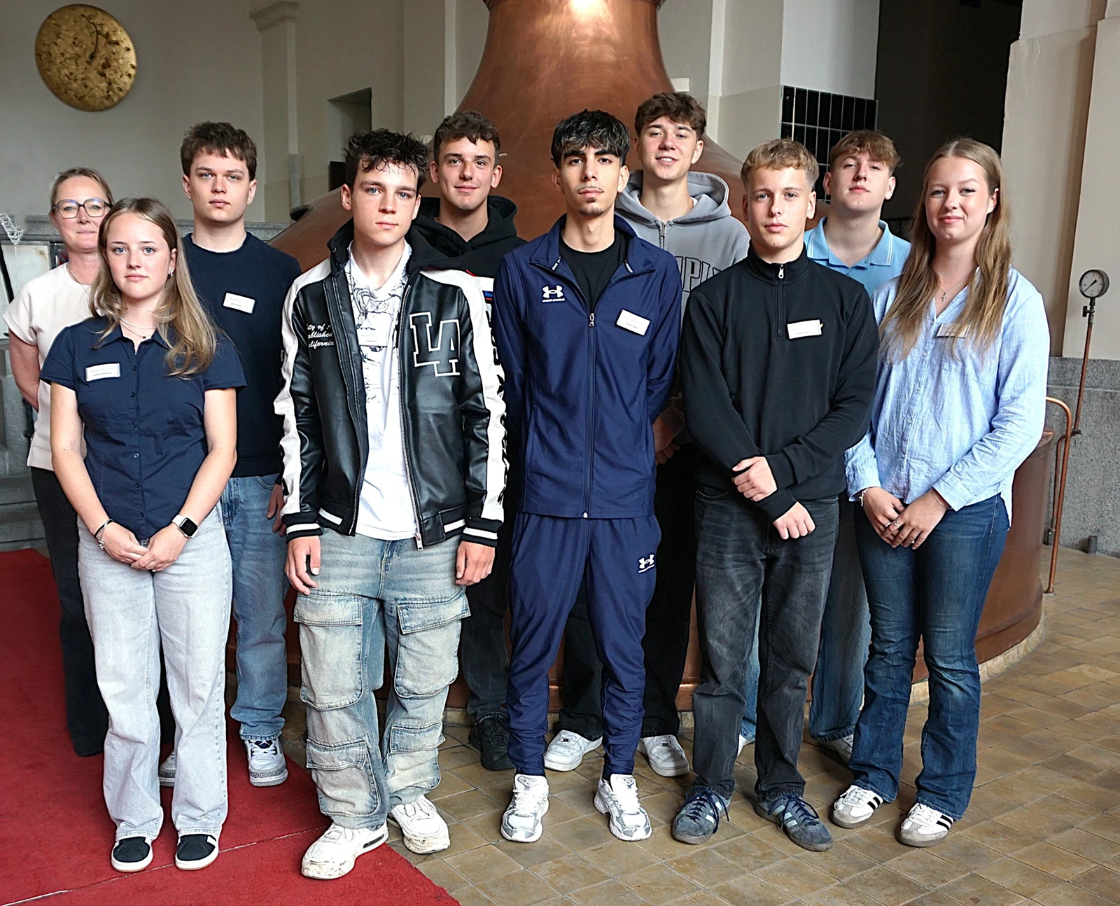 Eine Gruppe junger Menschen steht in zwei Reihen nebeneinander und blickt zur Kamera. Im Hintergrund ein Kupferkessel.