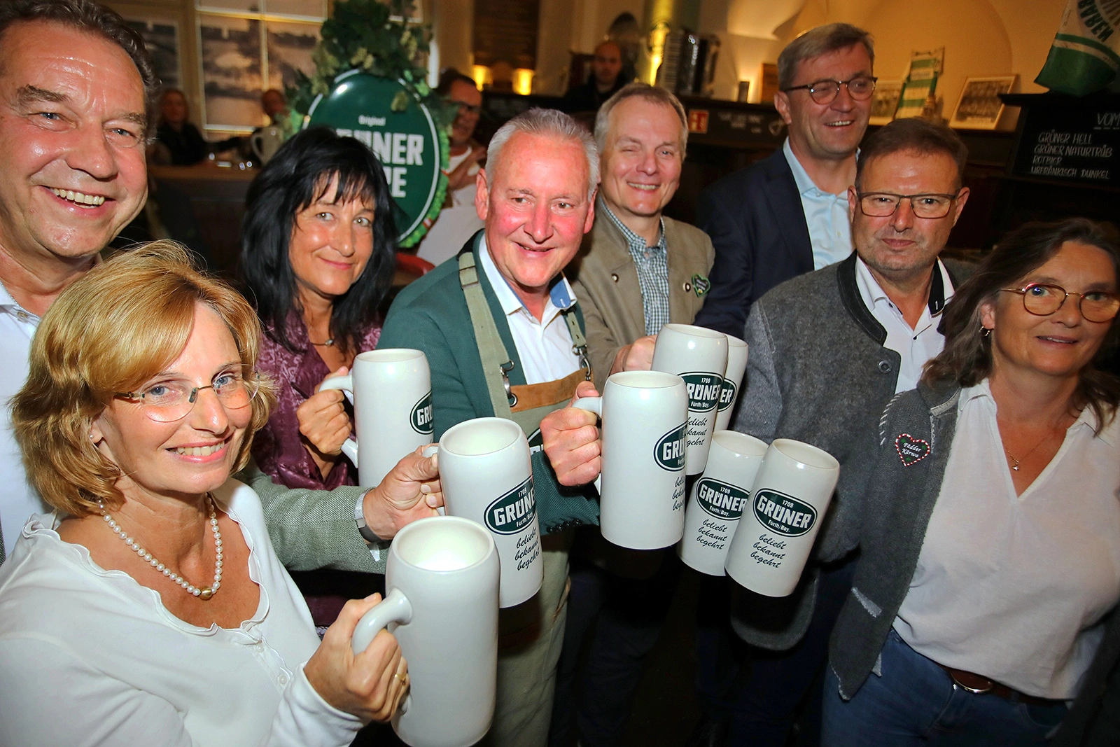 Mehrere Personen halten weiße Bierkrüge der Marke Grüner in der Hand. Im Hintergrund ist eine grün dekorierte Innenraumszene.