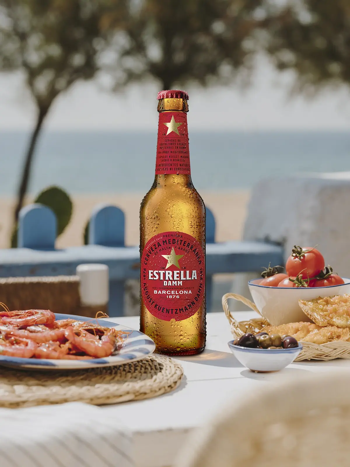 Eine Bierflasche der Marke Estrella Damm steht auf einem gedeckten Tisch. Im Hintergrund sind Meer, Strand und Bäume.