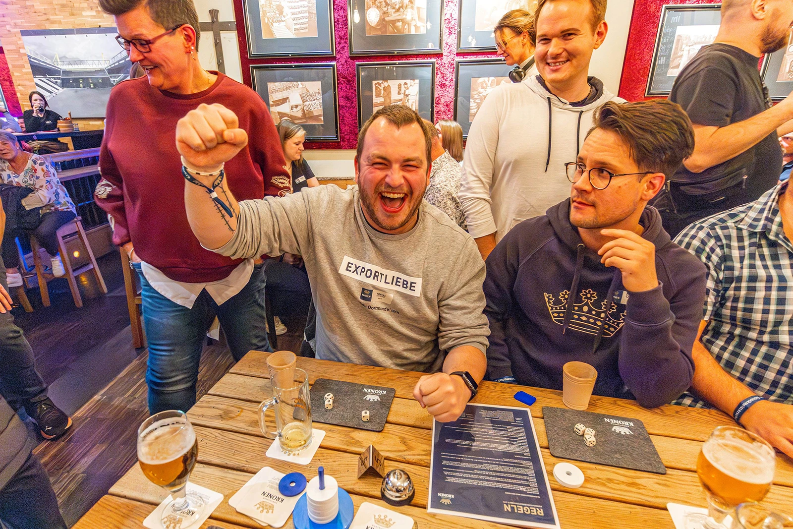Mehrere Personen sitzen an einem Holztisch mit Würfeln und Biergläsern. Eine Person jubelt, während andere lächeln.