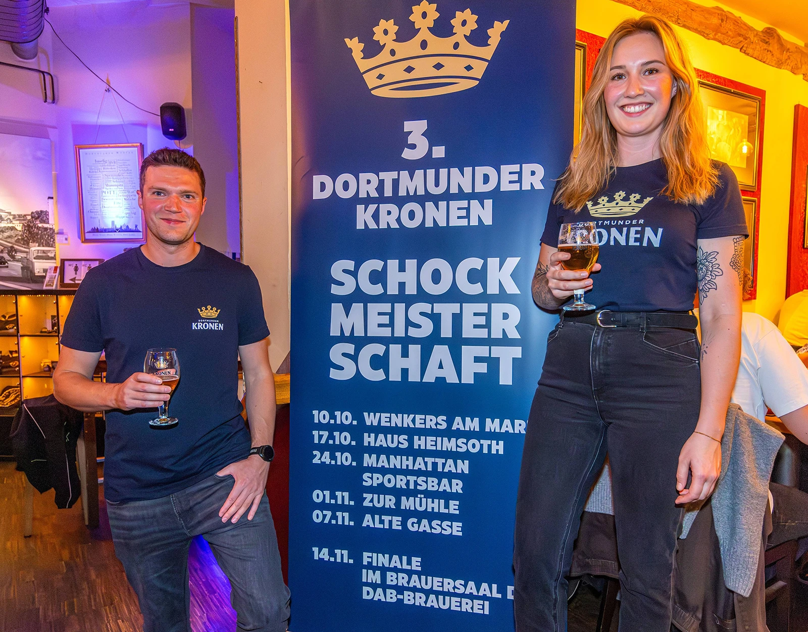Zwei Personen stehen vor einem Schockmeisterschaft Banner und halten jeweils ein Glas Dortmunder Kronen Bier in der Hand.