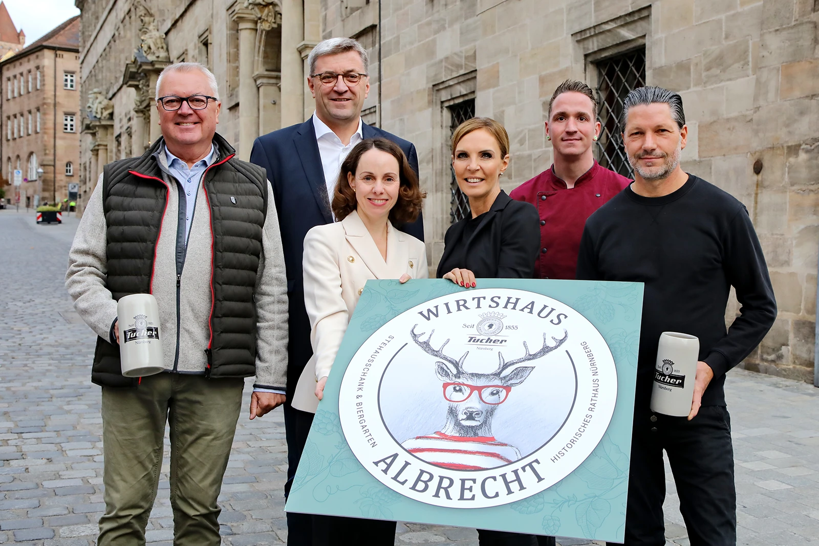 Sechs Personen halten ein Schild mit der Aufschrift Wirtshaus Albrecht. Im Hintergrund eine historische Sandsteinfassade.