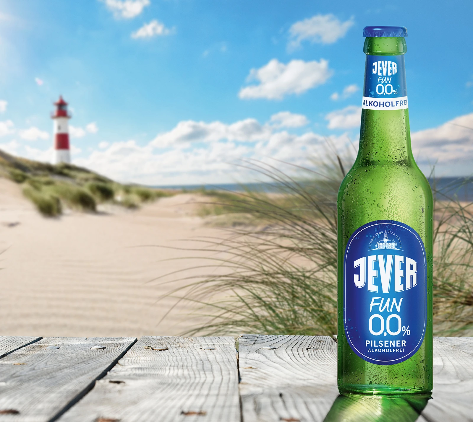 Eine grüne Flasche Jever Fun 0,0 Prozent steht auf Holzplanken. Im Hintergrund Sanddünen und ein roter Leuchtturm.