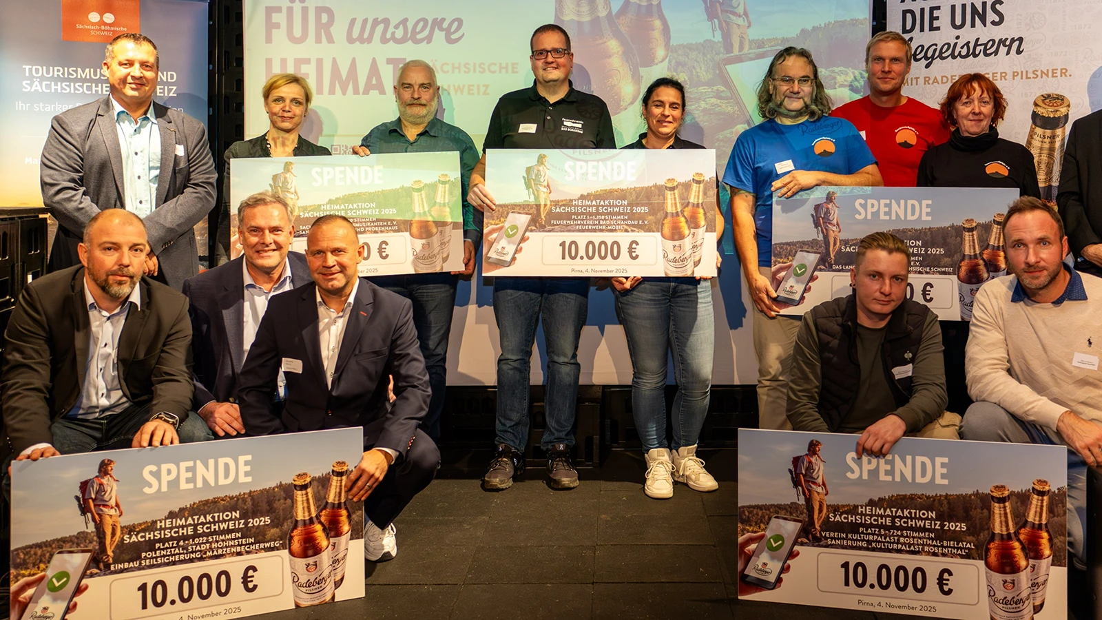 Mehrere Personen posieren mit großen Spendenschecks. Im Hintergrund Banner zur Heimataktion von Radeberger und der Sächsischen Schweiz.