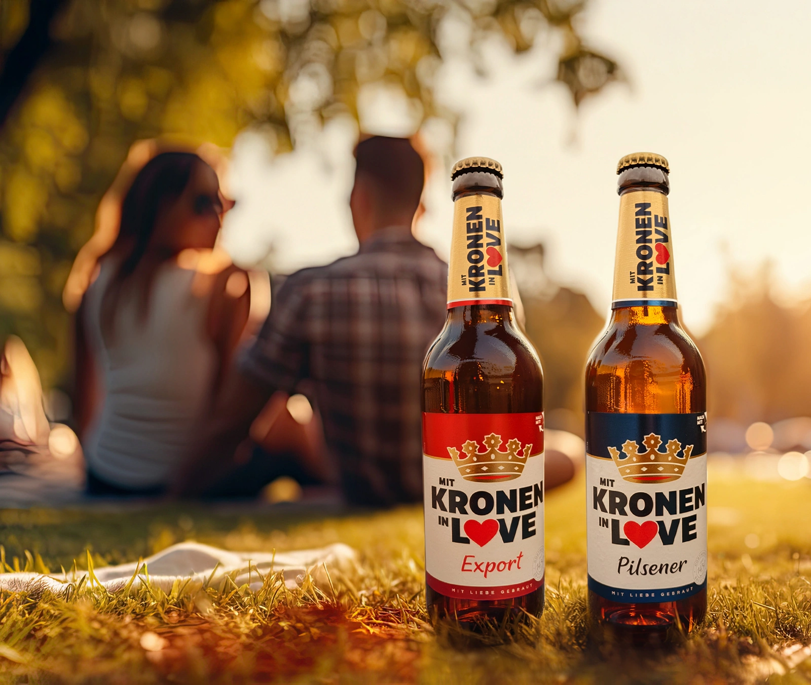 Zwei Dortmunder Kronen Bierflaschen stehen im Gras im Vordergrund. Im Hintergrund ein Paar im Park bei Sonnenuntergang.