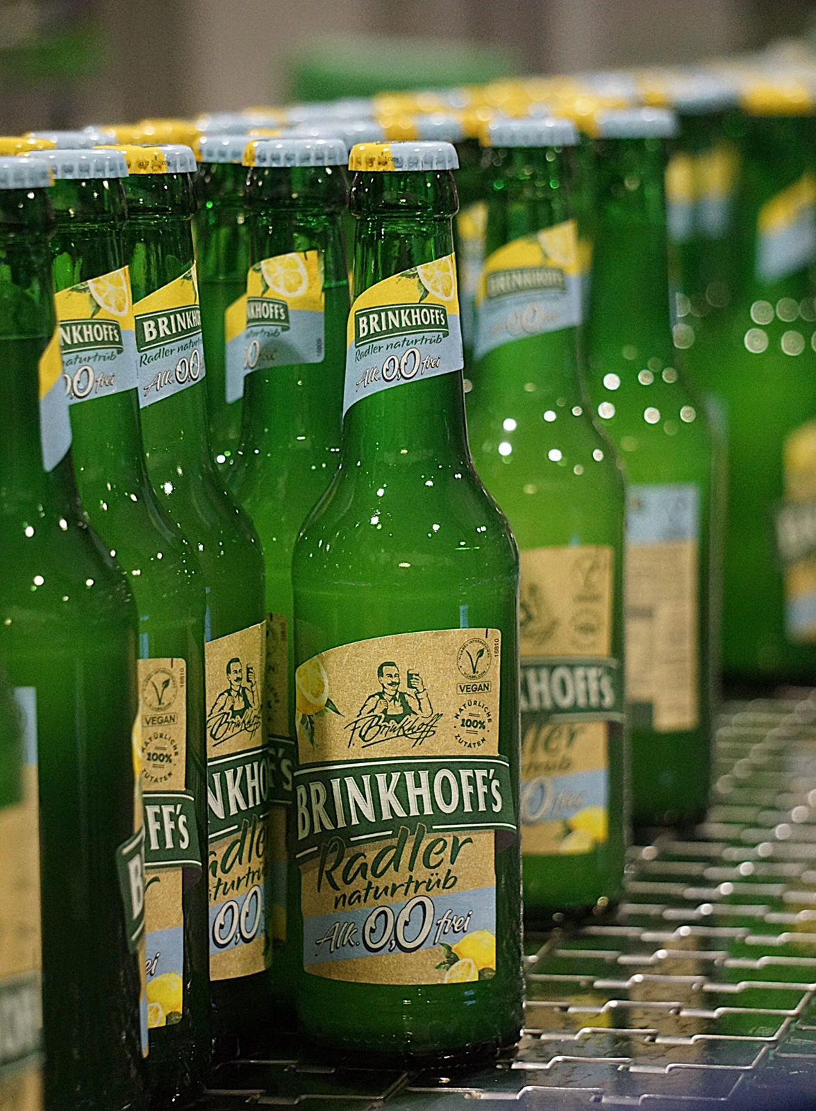 Mehrere Glasflaschen Brinkhoff’s Radler naturtrüb 0,0 stehen in Reihen auf einem Metallförderband.