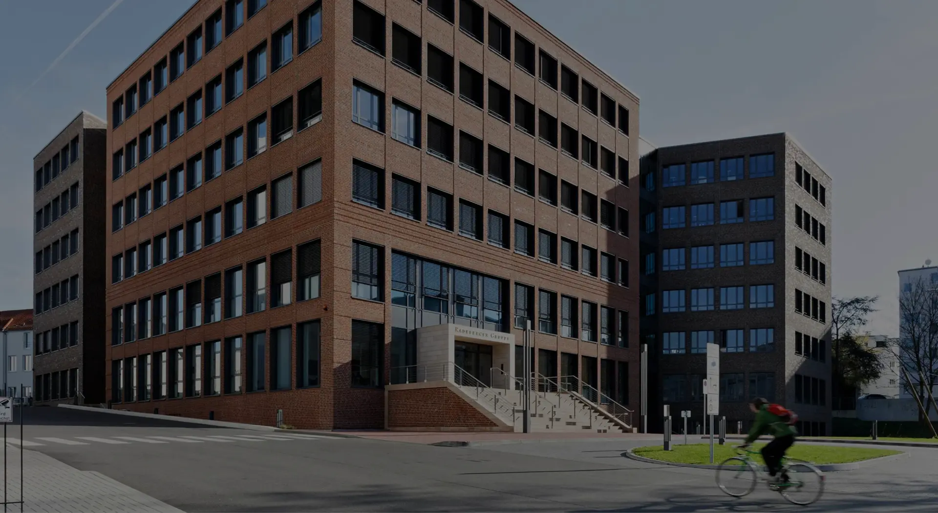 Ein modernes Bürogebäude aus Backstein mit vielen Fenstern steht an einer Straßenecke. Ein Radfahrer fährt vorbei.