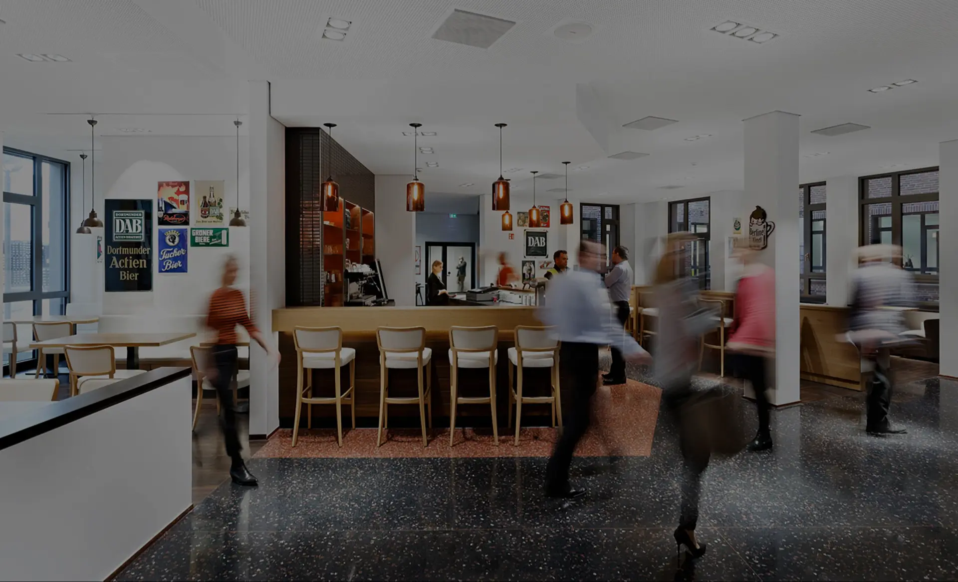 Menschen gehen durch eine moderne Bar mit Holztischen und Theke, beleuchtet durch Hängelampen.