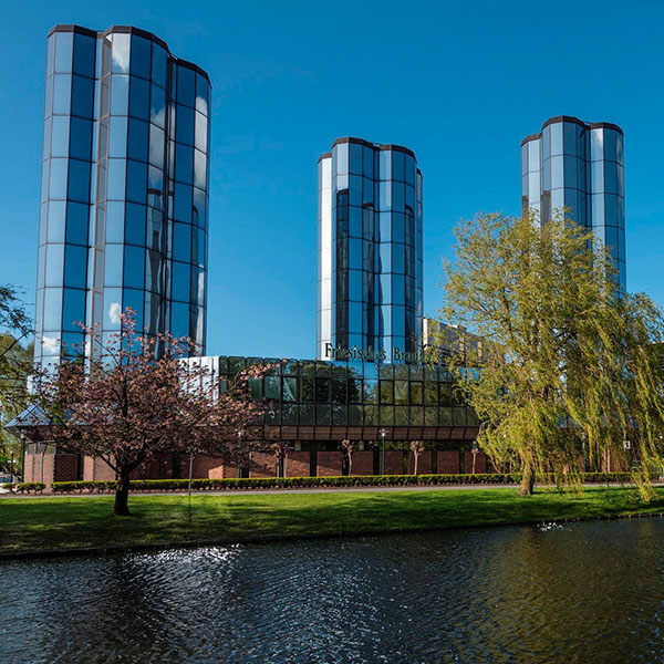 Drei hohe, gläserne Gebäude stehen nebeneinander. Ein Kanal und grüne Vegetation umgeben die Architektur bei sonnigem Wetter.