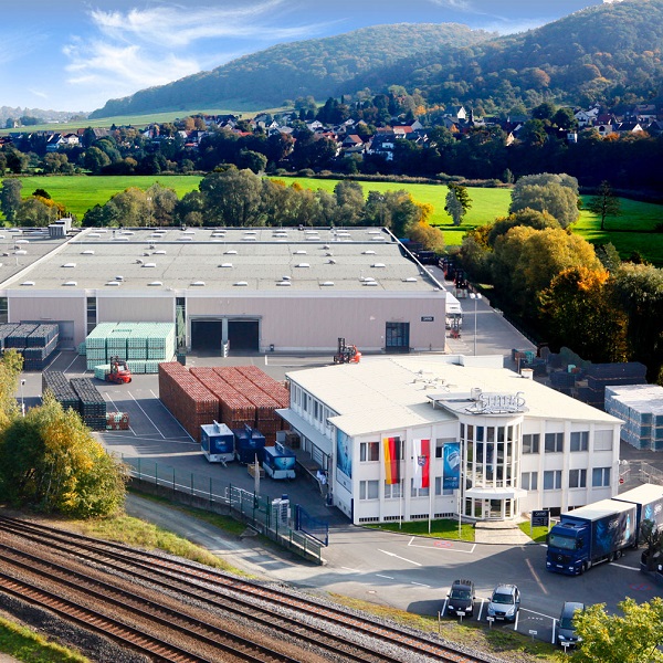 Luftaufnahme eines großen Industriegebäudes mit Bürotrakt und LKWs. Grüne Landschaft und Hügel im Hintergrund.