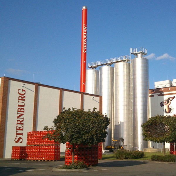 Eine Fabrik mit großen silberfarbenen Silos und einem hohen rotem Schornstein steht unter blauem Himmel.