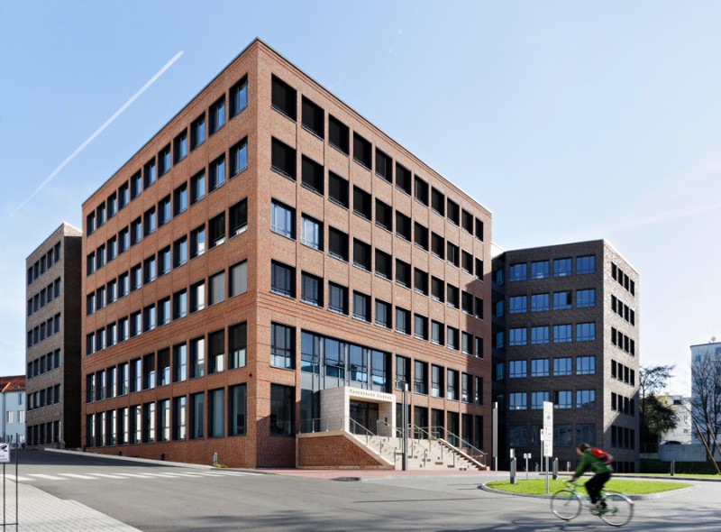 Ein modernes Bürogebäude aus rotem Backstein mit großen Fenstern, vor dem ein Radfahrer auf der Straße vorbeifährt.