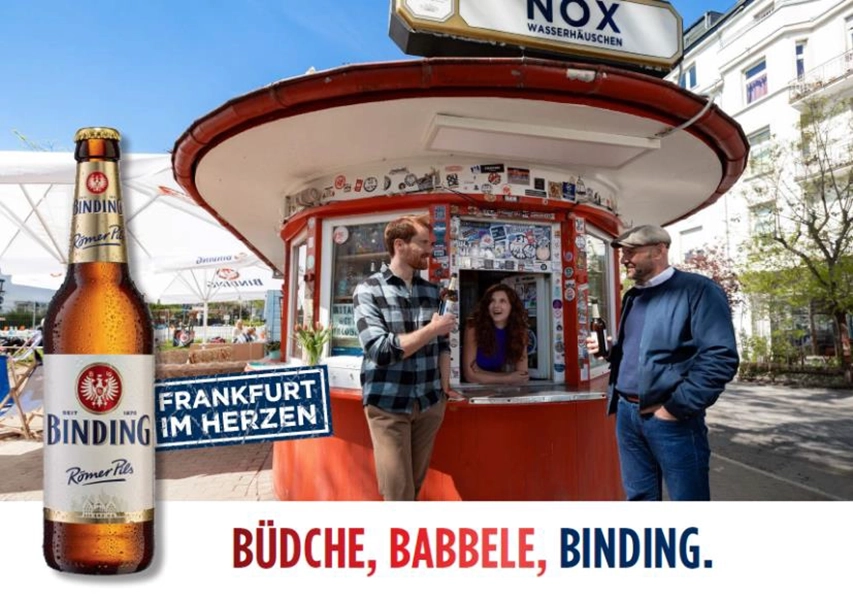 Zwei Männer stehen bei einem runden Kiosk und halten Getränke, während sie sich unterhalten.