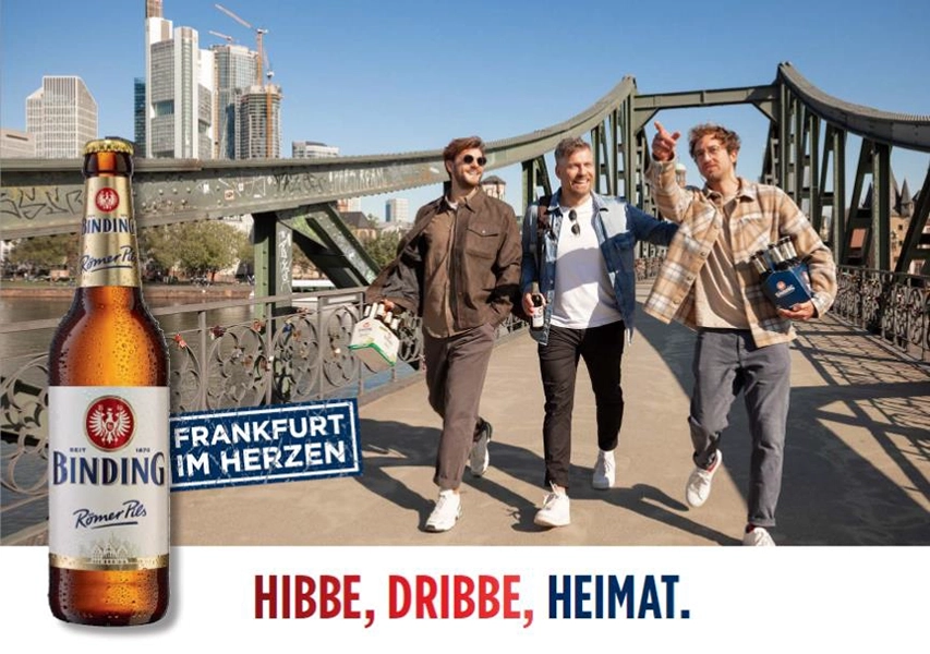 Drei Männer gehen lachend auf einer Brücke mit einer Großstadt im Hintergrund.