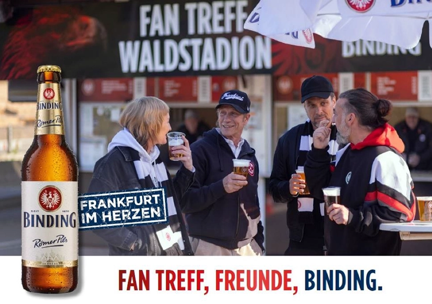 Drei Personen unterhalten sich im Freien an einem Getränkestand bei einem Sportevent.