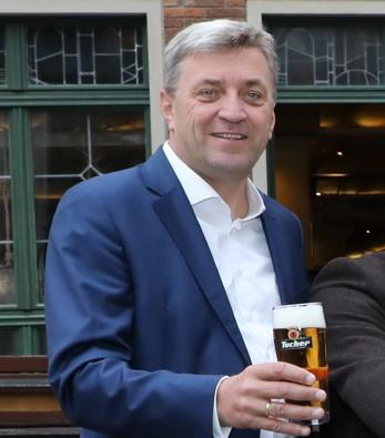 Ein Mann in einem blauen Anzug hält lächelnd ein Bierglas.