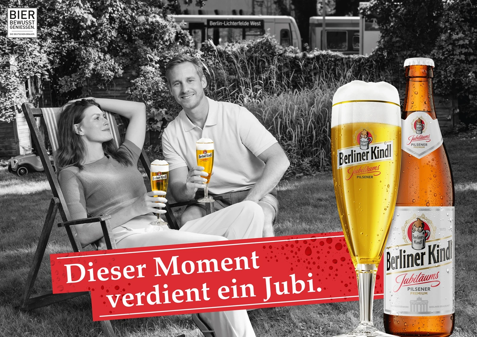 Ein Mann und eine Frau sitzen lächelnd auf Gartenstühlen und halten gefüllte Biergläser. Ein Zug fährt im Hintergrund vorbei.