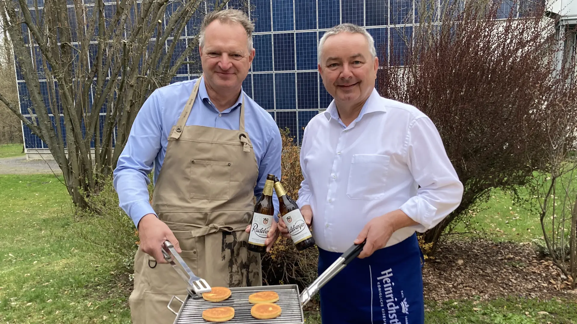 Zwei Männer stehen draußen und lächeln, einer grillt Pancakes und der andere hält Bierflaschen.