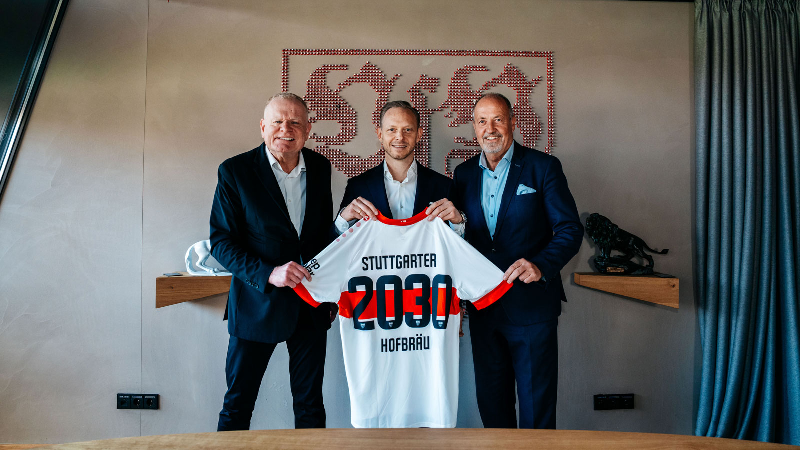 Drei Männer in Anzügen halten gemeinsam ein Fußballtrikot in einem Raum. Das Trikot zeigt die Jahreszahl 2030.