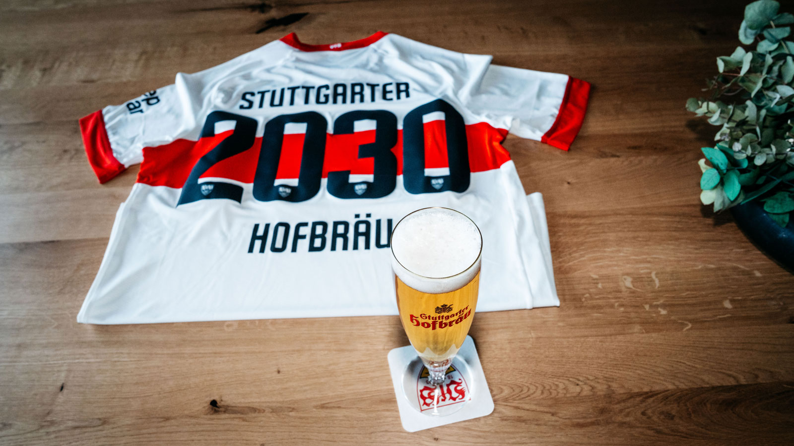 Ein Trikot mit der Aufschrift 2030 liegt auf einem Holzboden neben einem vollen Bierglas.