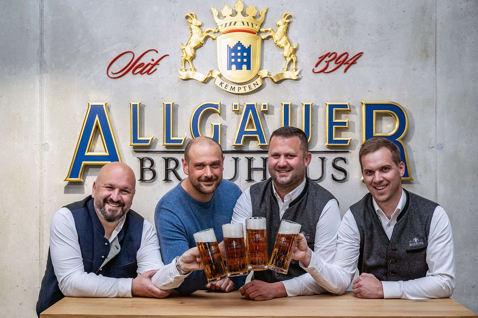 Vier Männer in traditioneller Kleidung stoßen mit Biergläsern an, im Hintergrund ein Brauerei-Logo.