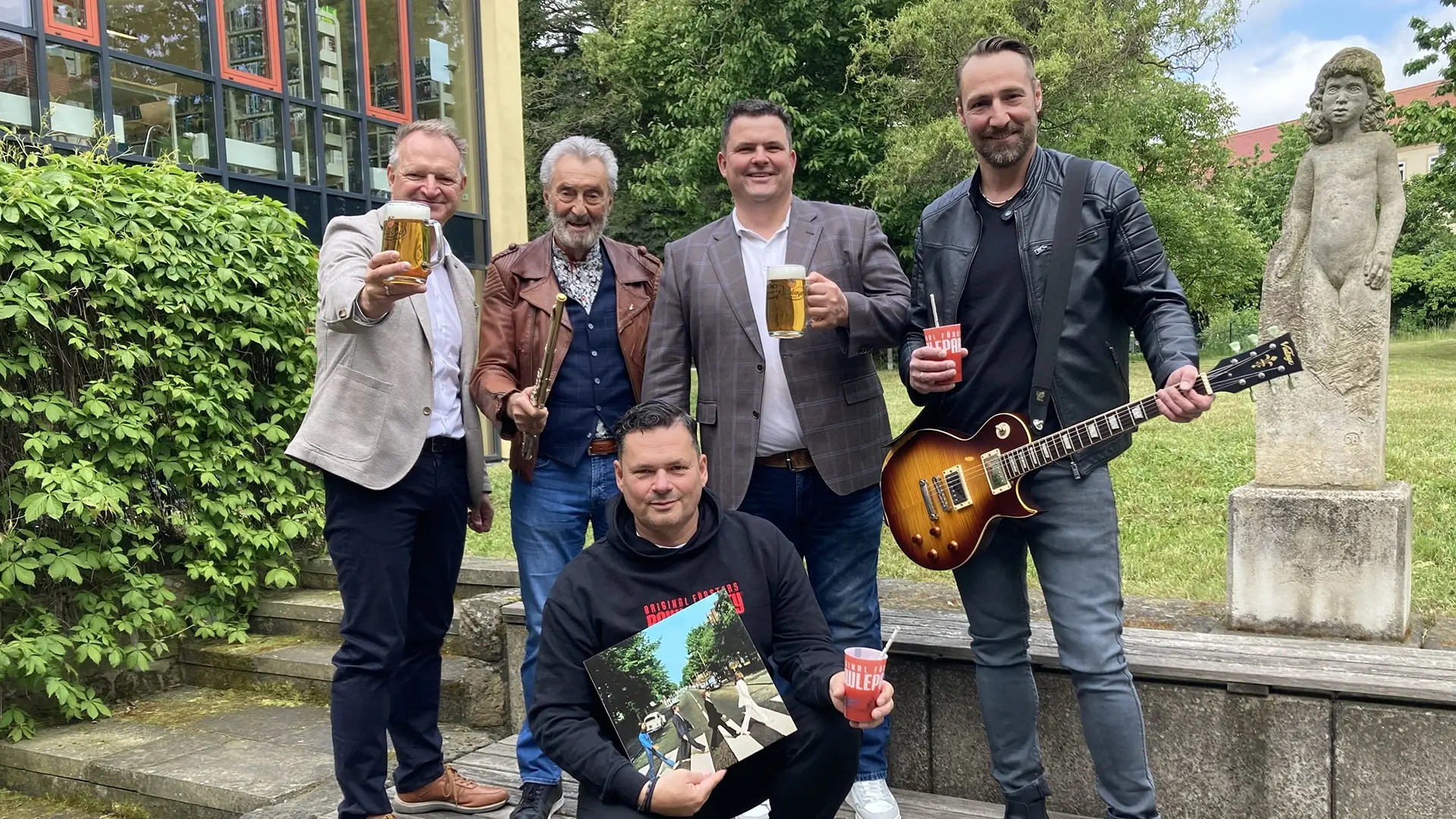 Fünf Männer stehen lächelnd mit Getränken und einer Gitarre im Freien vor einer Statue.