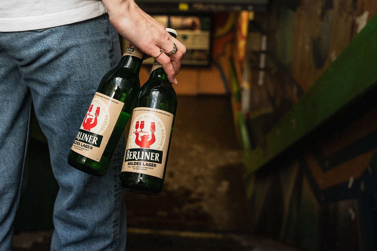 Person hält in einer Hand zwei Bierflaschen Berliner Mildes Lager. Im Hintergrund ein schmaler Durchgang mit Wandgraffiti.