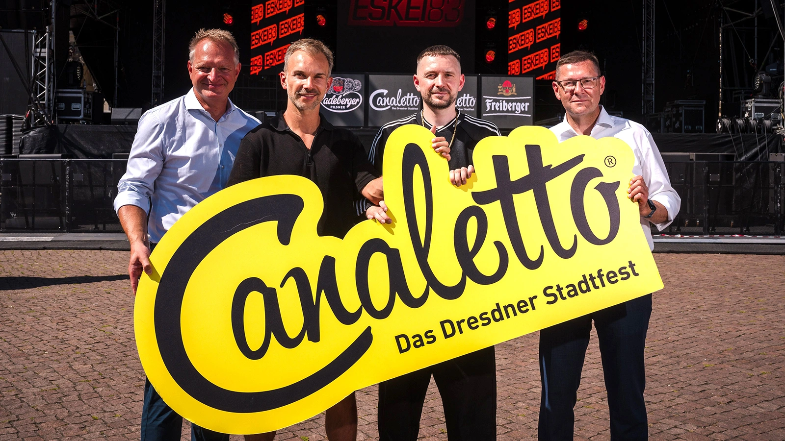 Vier Männer halten ein großes gelbes Schild mit der Aufschrift Canaletto - Das Dresdner Stadtfest vor einer Bühne.