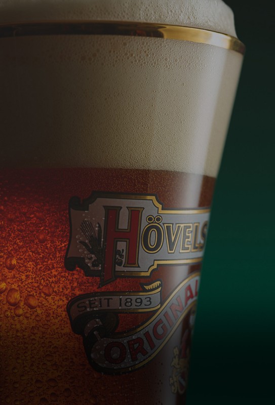 Nahaufnahme eines Bierglases mit Schaum und einem Etikett, vor einem dunklen Hintergrund.