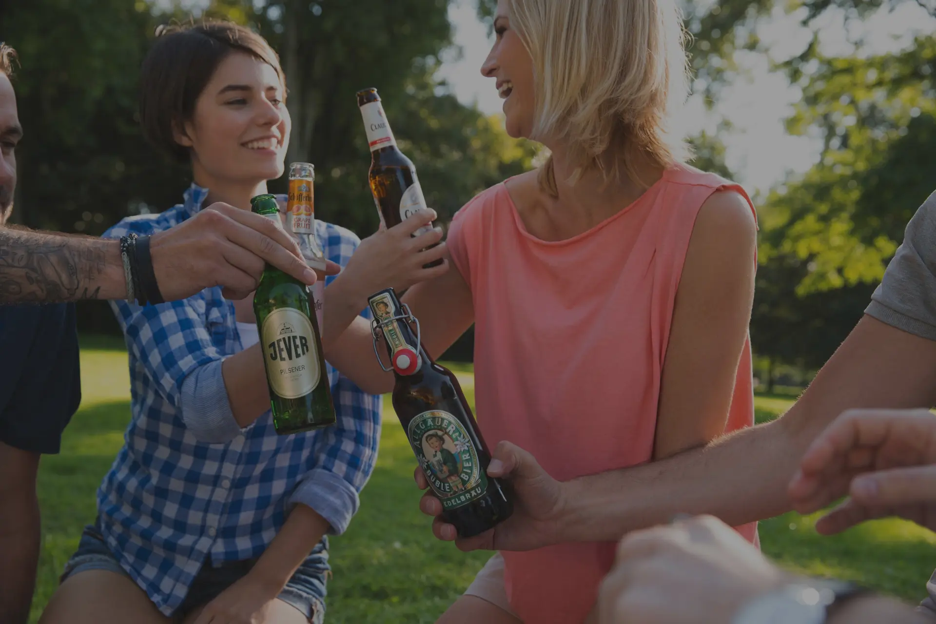 Vier Freunde stoßen im Park mit Flaschenbier an, lächeln und genießen die Sonne.